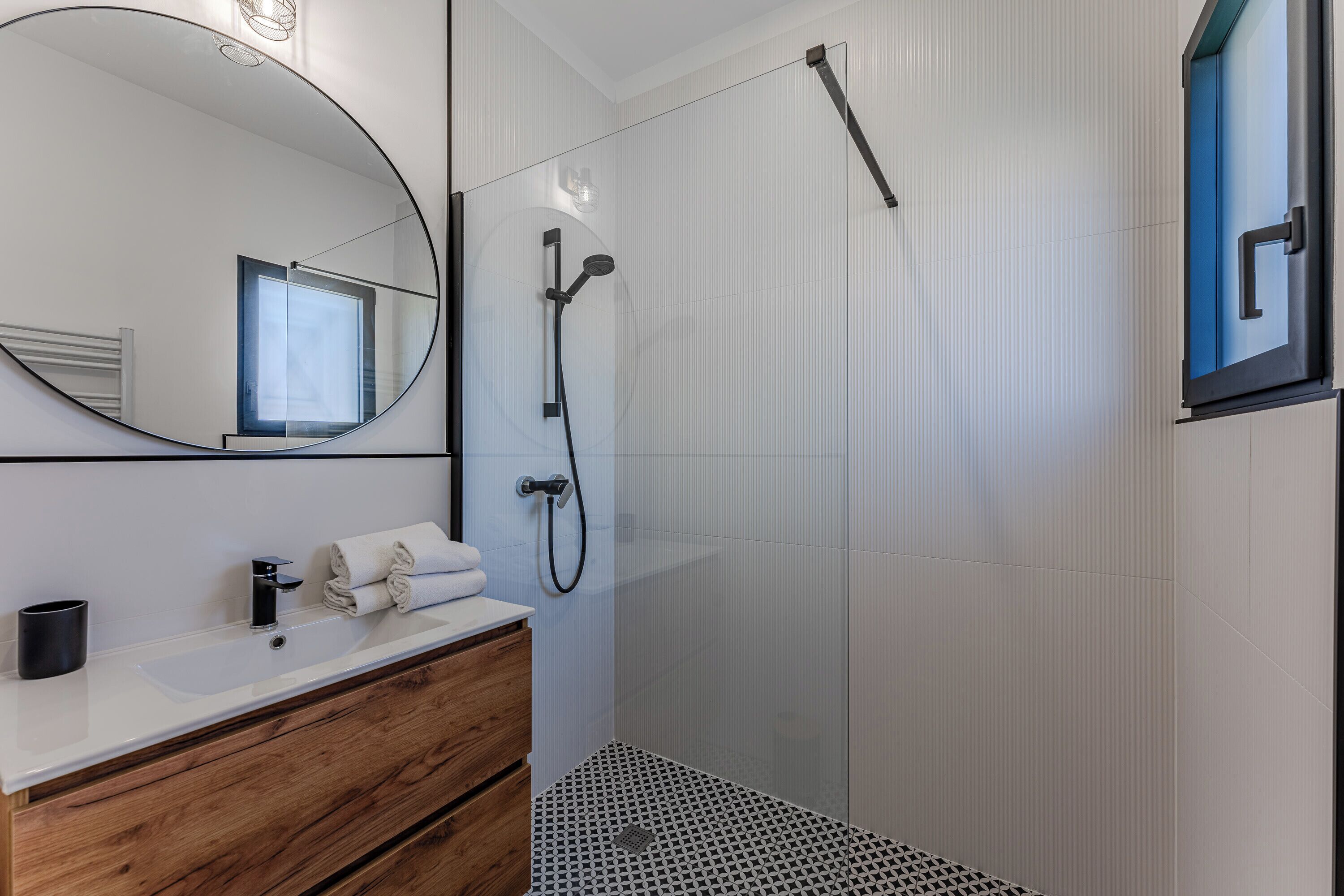 A modern bathroom with a wooden vanity, a large mirror, a shower enclosure, and a patterned floor.