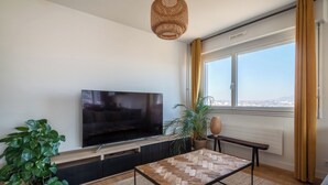 A modern living room with a flat-screen TV, a wooden coffee table, a large potted plant, and a view through a window. - Appartement Mi-Luna - Welkeys (Tassin-la-Demi-Lune)