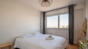 A neatly made bed with white linens, a bedside table, and a window with curtains. - Appartement Mi-Luna - Welkeys (Tassin-la-Demi-Lune)