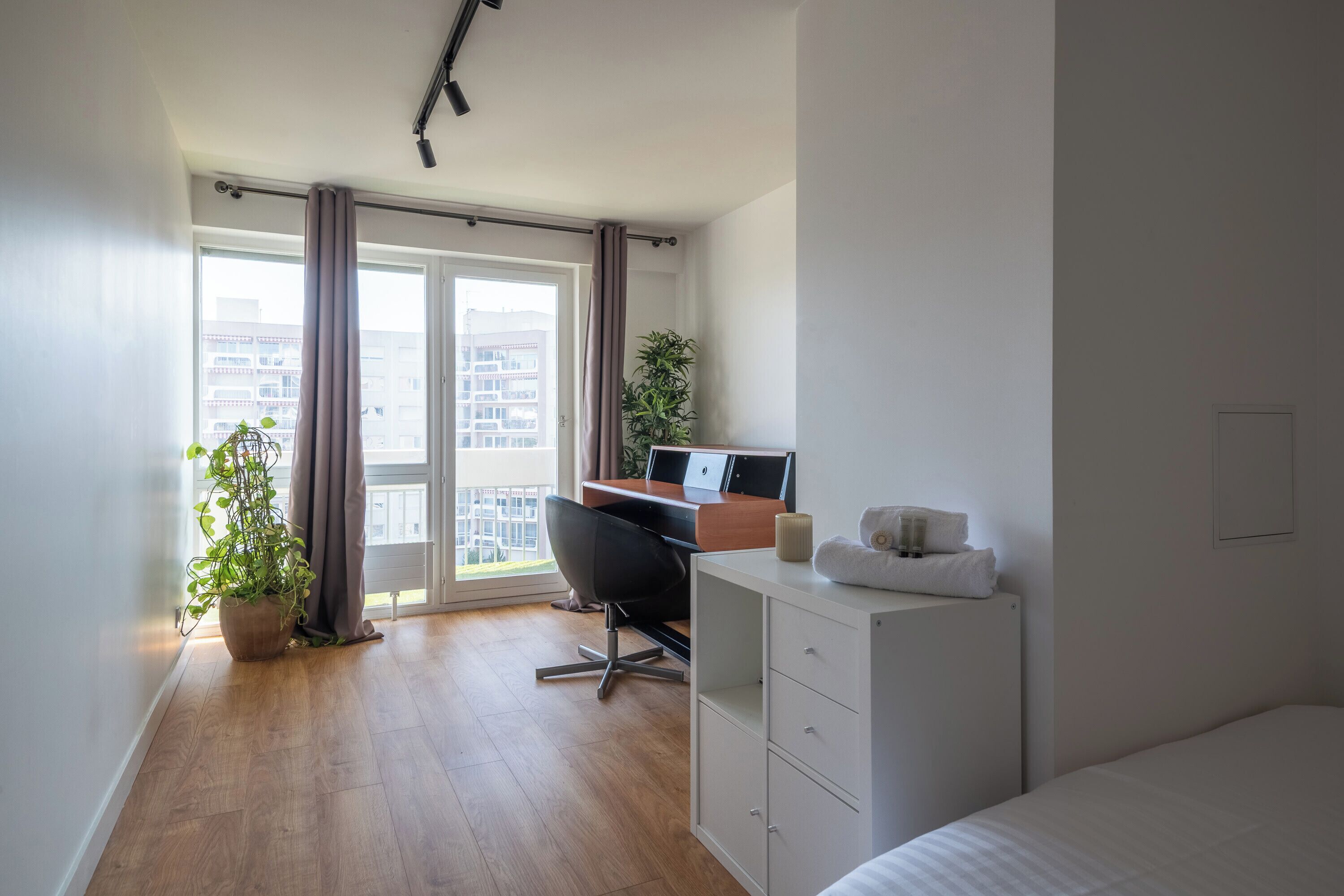 Un dormitorio moderno con suelo de madera, un escritorio con computadora, una cama, una planta en maceta y vistas a edificios a través de un ventanal grande.