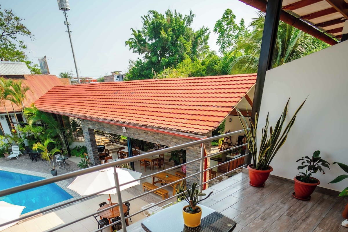 Uma área de estar coberta ao ar livre com móveis de vime e uma mesa, com vista para uma piscina e um edifício com telhado vermelho.