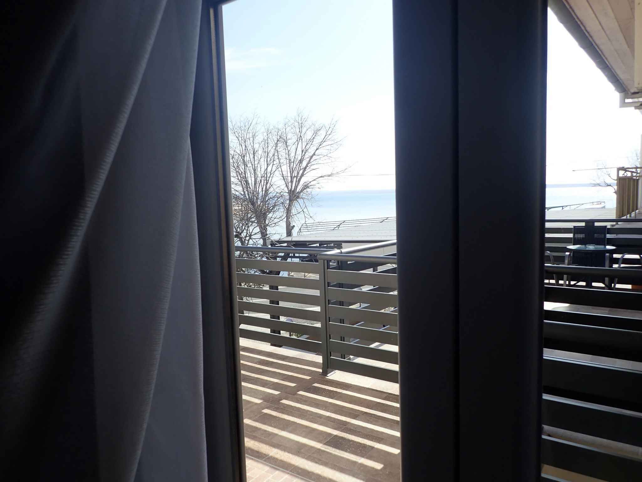 Une vue depuis une fenêtre ornée d’une rambarde en métal, donnant sur une zone pavée avec des tables et des chaises, et sur une étendue d’eau au loin.