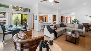 A spacious living area with a dining table, a sofa, and a coffee table. There is a kitchen in the background with a refrigerator and a sink. - Kapalua Ridge Villas 1023 (Lahaina)
