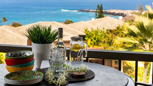 A table with colorful bowls, a bottle, a pitcher, and a potted plant, overlooking a coastal view with a thatched roof structure. - Kapalua Ridge Villas 1023 (Lahaina)
