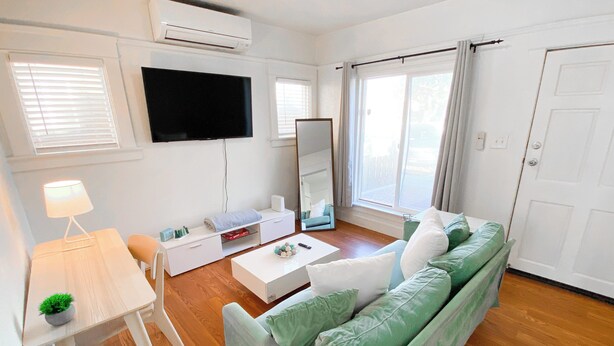 Een moderne woonkamer met een groene bank, een witte salontafel, een flatscreen-televisie aan de muur en een bijzettafeltje met een lamp en een plant.