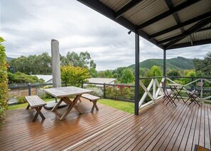 Una terraza techada al aire libre con piso de madera, mesas de picnic y vistas a un paisaje exuberante con colinas y árboles.