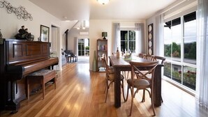 A spacious living room with a piano, dining table, and chairs. There is a bookshelf and a large window with a view of greenery.