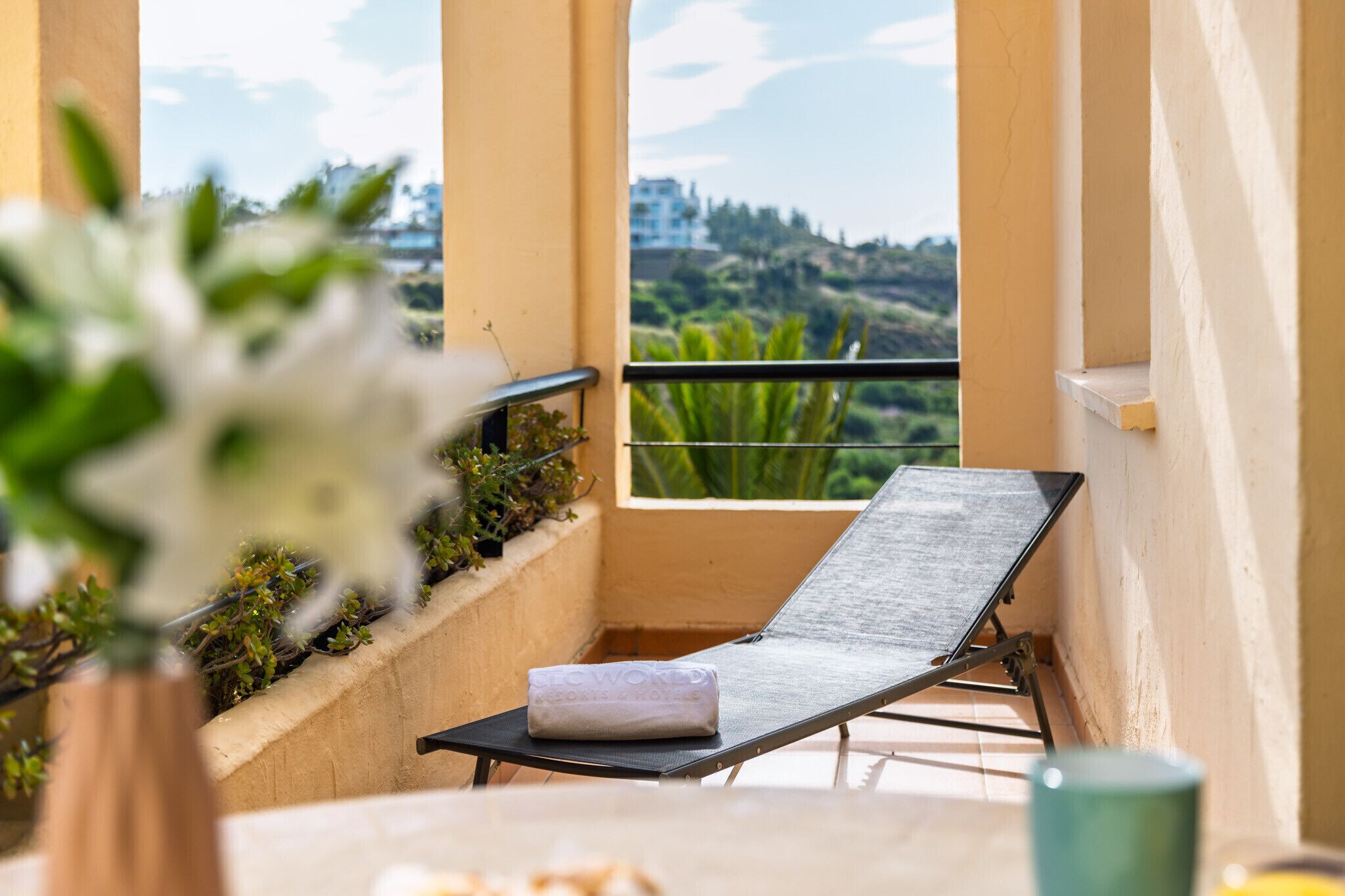A balcony with a sun lounger, a towel, and a view of greenery and buildings.