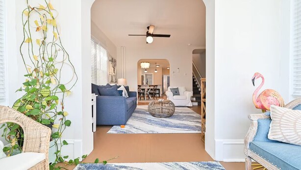 Un salon moderne avec un canapé bleu, un fauteuil blanc et une chaise en osier. Une grande plante se trouve à gauche, et une décoration représentant un flamant rose est placée à droite.
