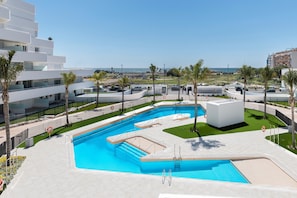 A modern pool area with a clear view of the sea, surrounded by white buildings and palm trees.