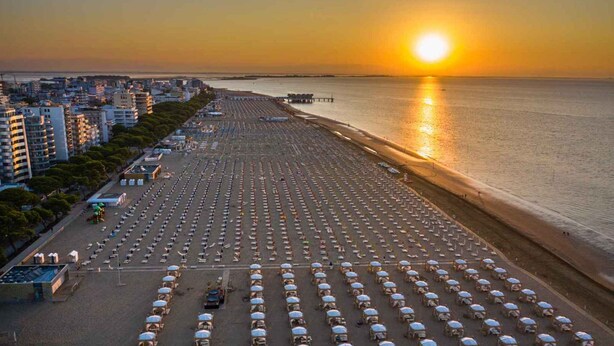 A beach with rows of umbrellas and sun loungers, a pier in the background, and buildings along the coastline. - Holiday apartment with air conditioning and balcony (Lignano)