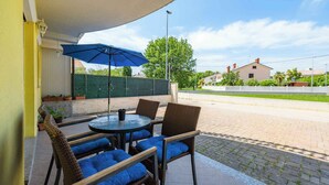 Une terrasse extérieure avec des meubles en osier et un parasol bleu, un chemin pavé et une vue sur des maisons en arrière-plan.
