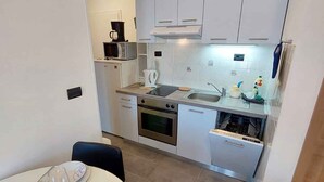 Une cuisine compacte avec des meubles blancs, un four à micro-ondes, une cuisinière et un évier. Un coin repas est aménagé avec une table pour deux personnes.