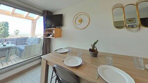 A dining area with a wooden table set for two, a television mounted on the wall, and a view of the outdoors.