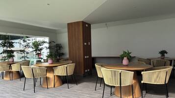 A modern indoor dining area with round wooden tables and woven chairs, surrounded by vertical wooden panels and potted plants.