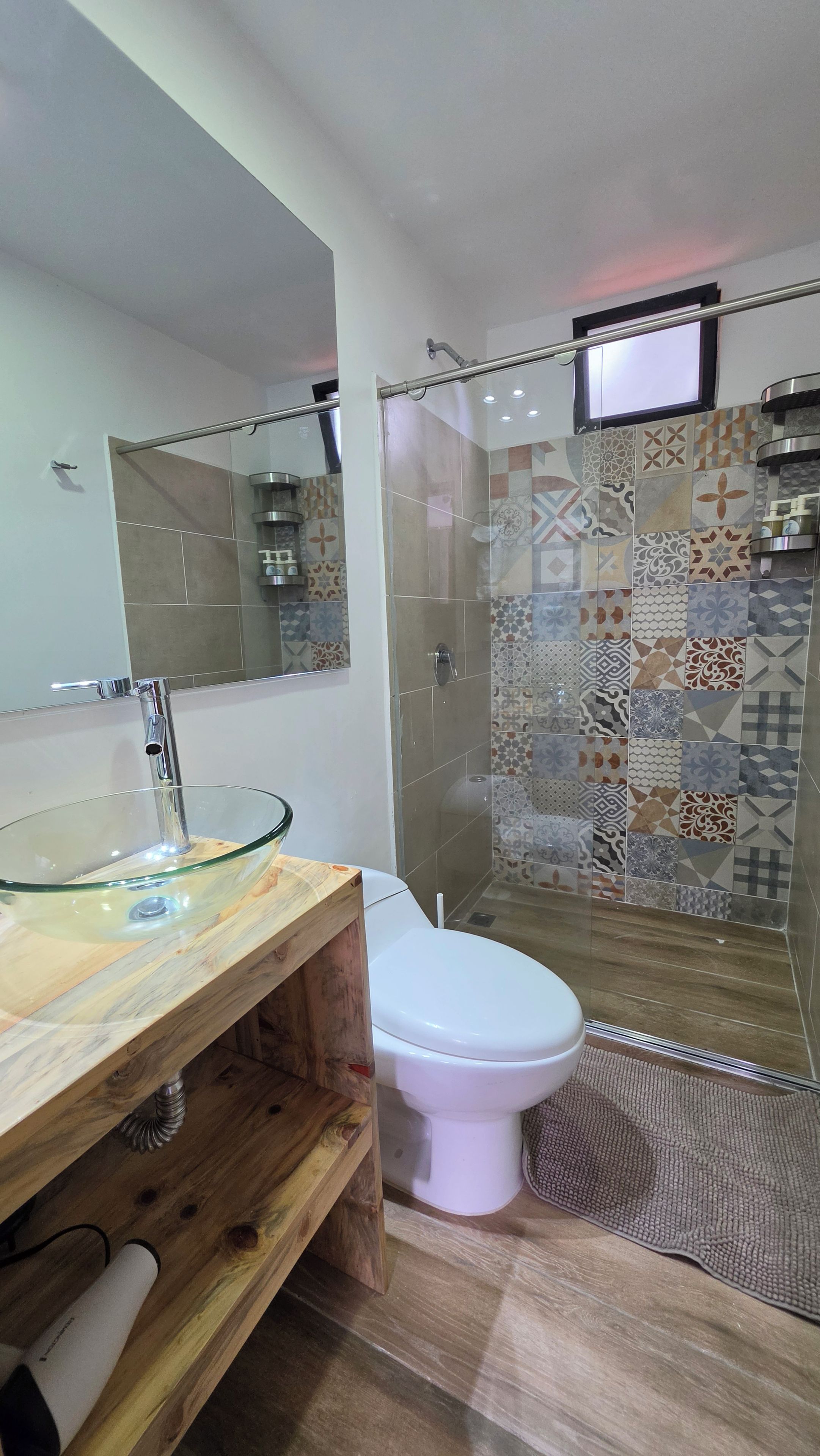 Un baño con lavamanos de vidrio, inodoro blanco y ducha con azulejos estampados.