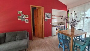 A room with a red wall, a wooden door, a grey sofa, a wooden dining table with blue chairs, and a kitchen area visible through an open door. - Hotel La Caracola Salento (Salento)