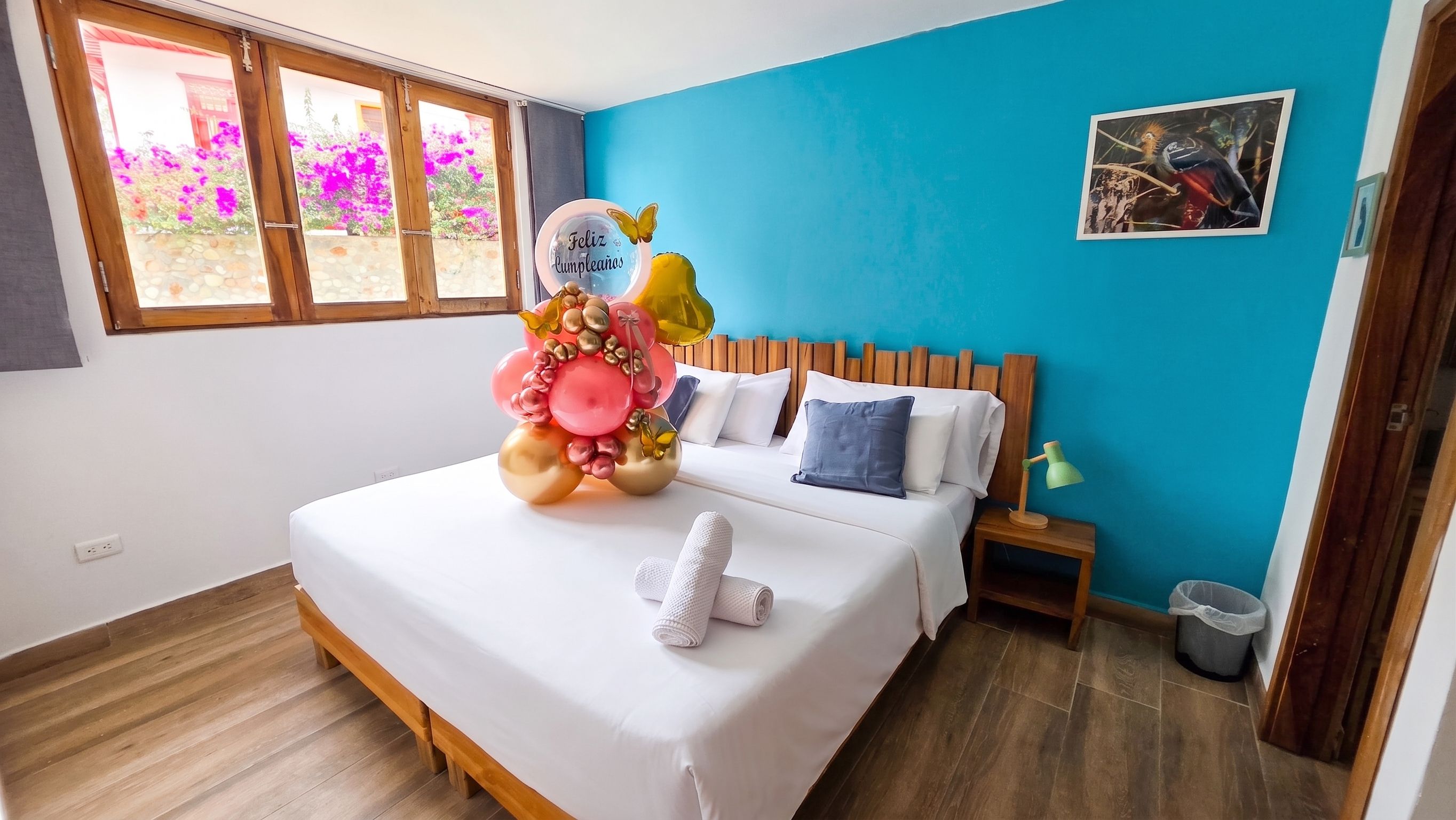 Un dormitorio con cama, almohadas, una mesita de noche, lámpara, un cubo de basura y una ventana con vistas a flores.