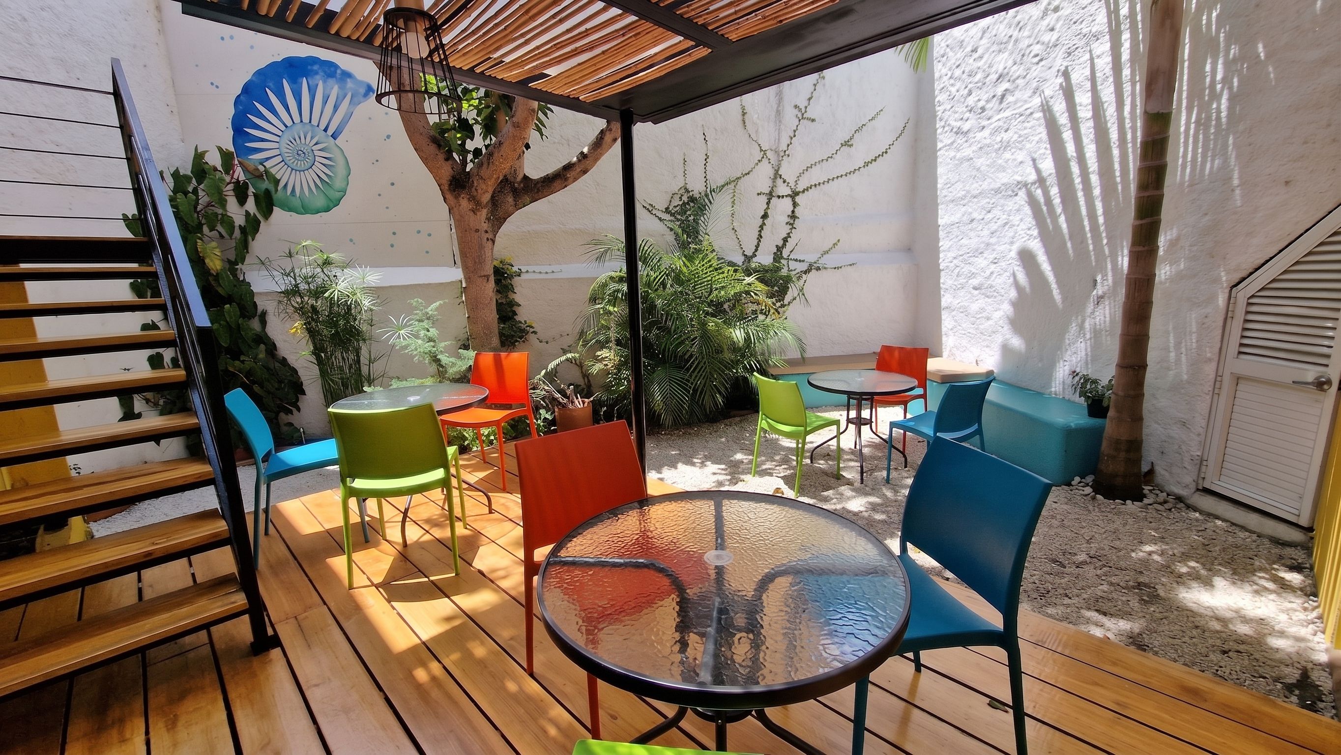 Un patio exterior con piso de madera, una mesa de vidrio y sillas coloridas.