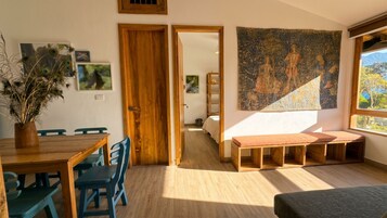 A room with a wooden dining table, chairs, a bench, a tapestry, and a view through a doorway.
