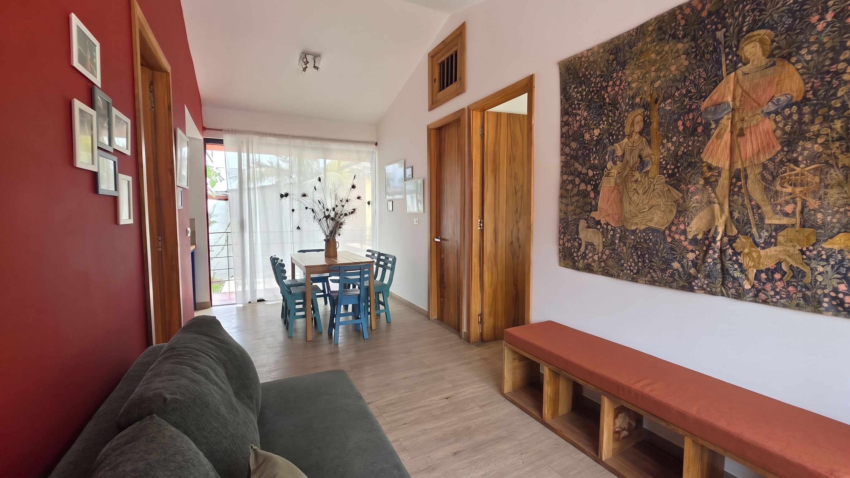 A modern living room with a sofa, a wooden bench, a dining table with chairs, and a large tapestry on the wall.