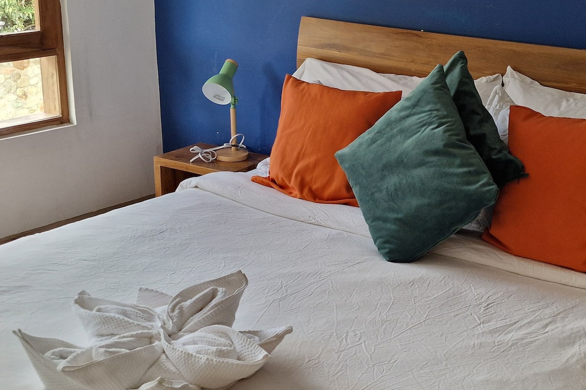A bed with white bedding, orange and green pillows, a wooden headboard, and a nightstand with a green lamp.