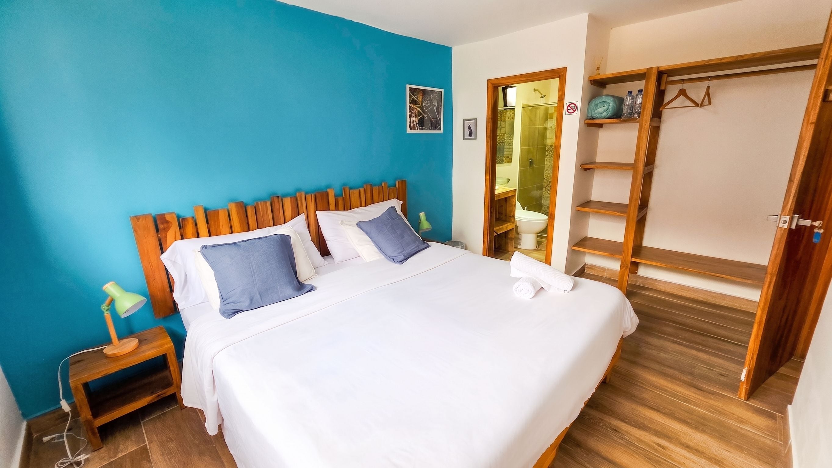 A hotel room with a bed, a nightstand, a lamp, a wooden headboard, a blue wall, a bathroom with a shower, and a wooden wardrobe.