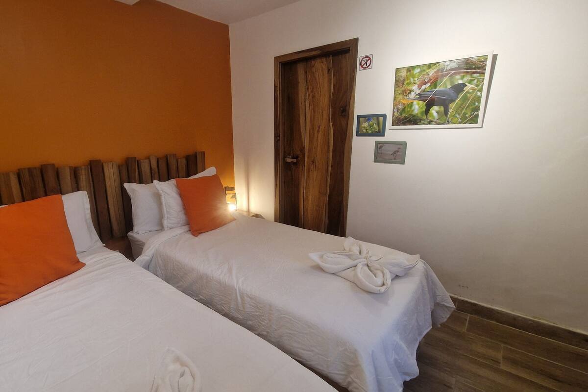 A room with two beds, each with white bedding and orange pillows, a wooden headboard, a wooden door, and a wall with framed pictures.