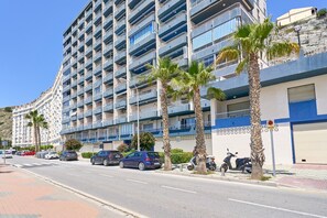Un edificio residencial de varios pisos con balcones, coches aparcados y palmeras a lo largo de la calle.
