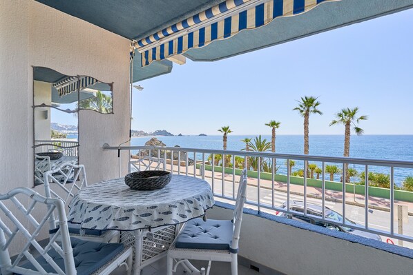 A balcony with a table and chairs, overlooking a coastal area with palm trees and the sea.