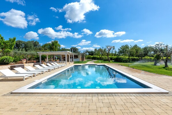 A rectangular swimming pool with lounge chairs and a covered seating area, surrounded by greenery and a clear sky.