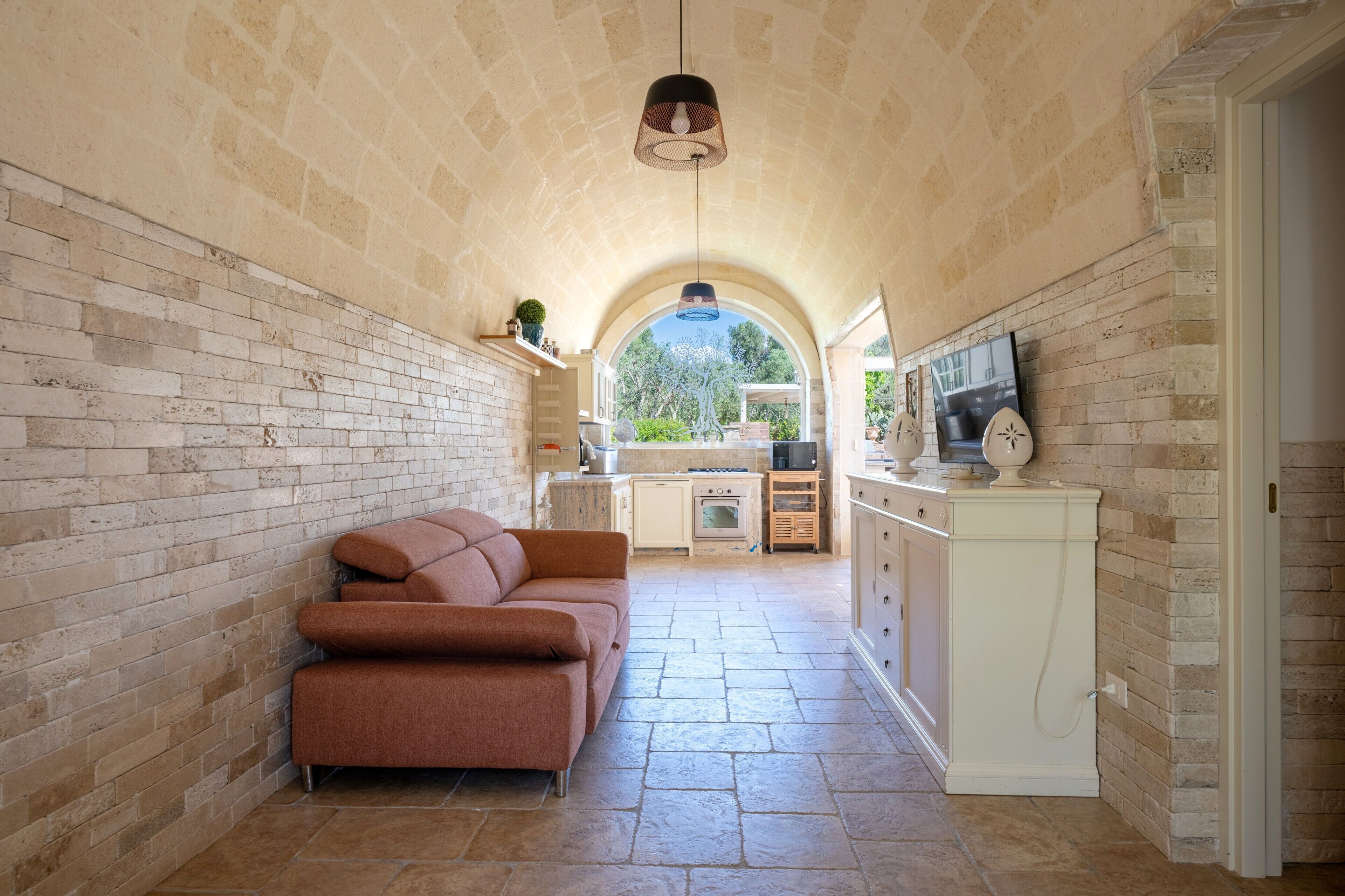 A narrow hallway with a brick wall, a sofa, a kitchen area with cabinets, and a television.