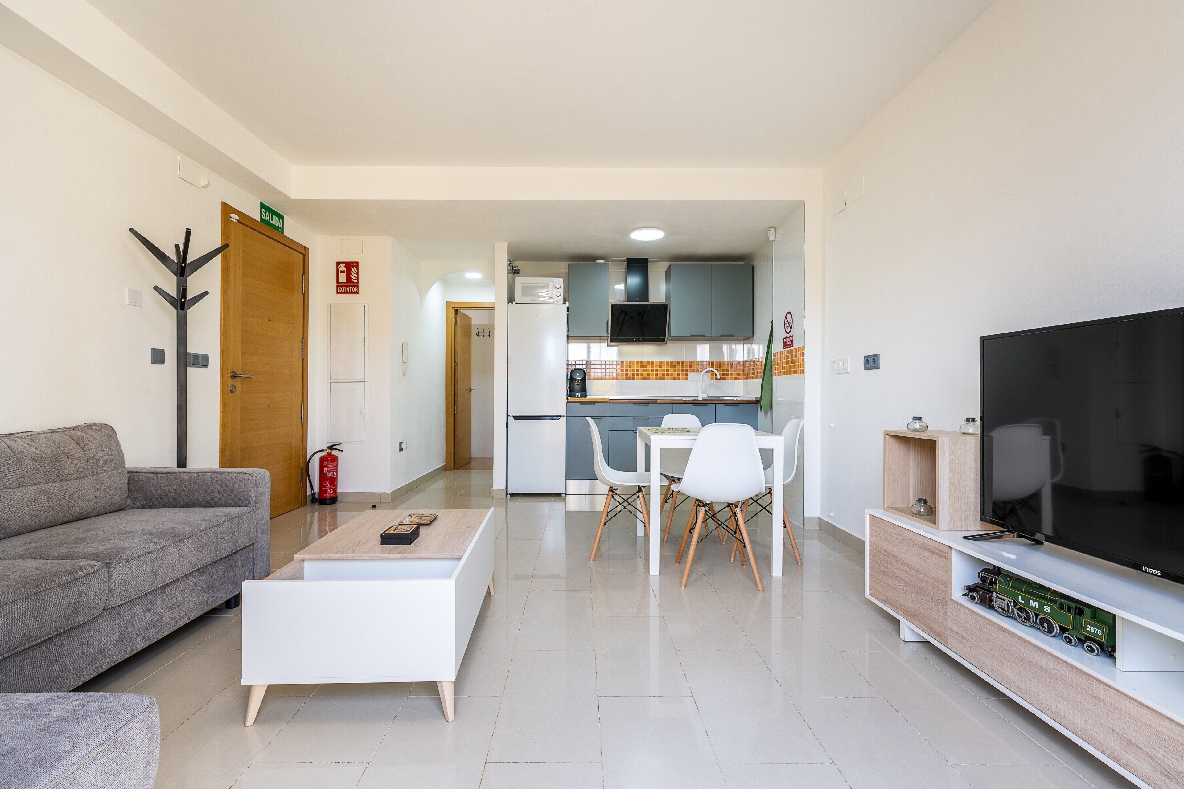 Una sala moderna con un sofá, una mesa de centro, televisión y una cocina al fondo.