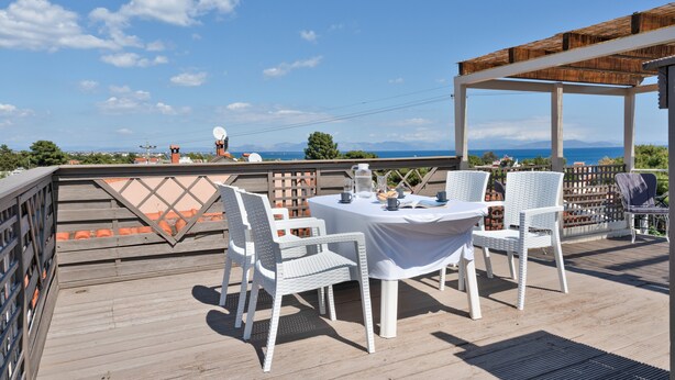 A deck with white wicker furniture, a table with a white tablecloth, and a view of the sea and mountains in the background.