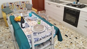 A dining area with a table set for two, a kitchen with a stove and oven, and a patterned sofa. - Apartment 'S'accomodassi - Tra Città E Mare' with Air Conditioning (Avola)