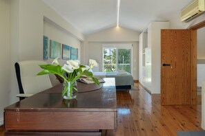 Une chambre avec un plancher en bois, un lit, un bureau agrémenté d’un vase de fleurs et une fenêtre donnant sur un beau paysage.