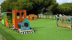 A playground with a train-themed play structure, a large tree, and a fenced area.
