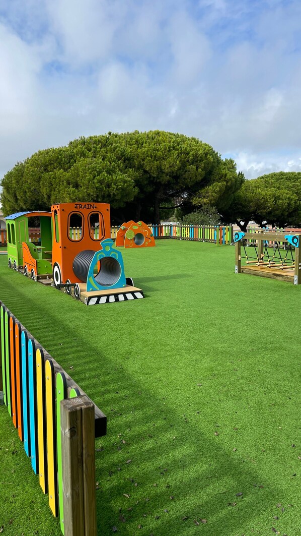Une aire de jeux avec une structure ludique sur le thème du train, un grand arbre et une zone délimitée par une clôture.