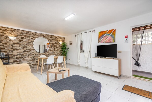 A living room with a sofa, a TV, a dining table with chairs, a plant, and a wall with a mirror and artwork.