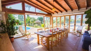 Un espacio interior amplio con una mesa de madera para cuatro personas, rodeada de sillas. Amplias ventanas ofrecen vistas al exterior, incluyendo una piscina y abundante vegetación.