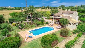 Una gran piscina con un sillón de sol y una estructura de juegos para niños, rodeada de exuberante vegetación y una casa de piedra.