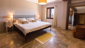 Un dormitorio con cama, mesitas de noche, lámpara, ventana con cortinas y una silla.