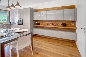 A modern kitchen with white cabinets, a wooden shelf, and a dining area with a glass table and chairs.