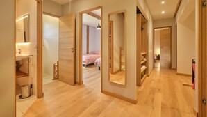 Een gang met houten vloer, openslaande deuren naar een slaapkamer met een bed en een badkamer met een wastafel en spiegel.