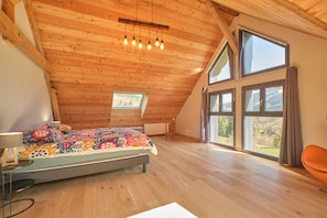 Un dormitorio con techo inclinado, una cama con colcha de colores, un escritorio con lámpara y ventanas amplias que ofrecen vistas al exterior.