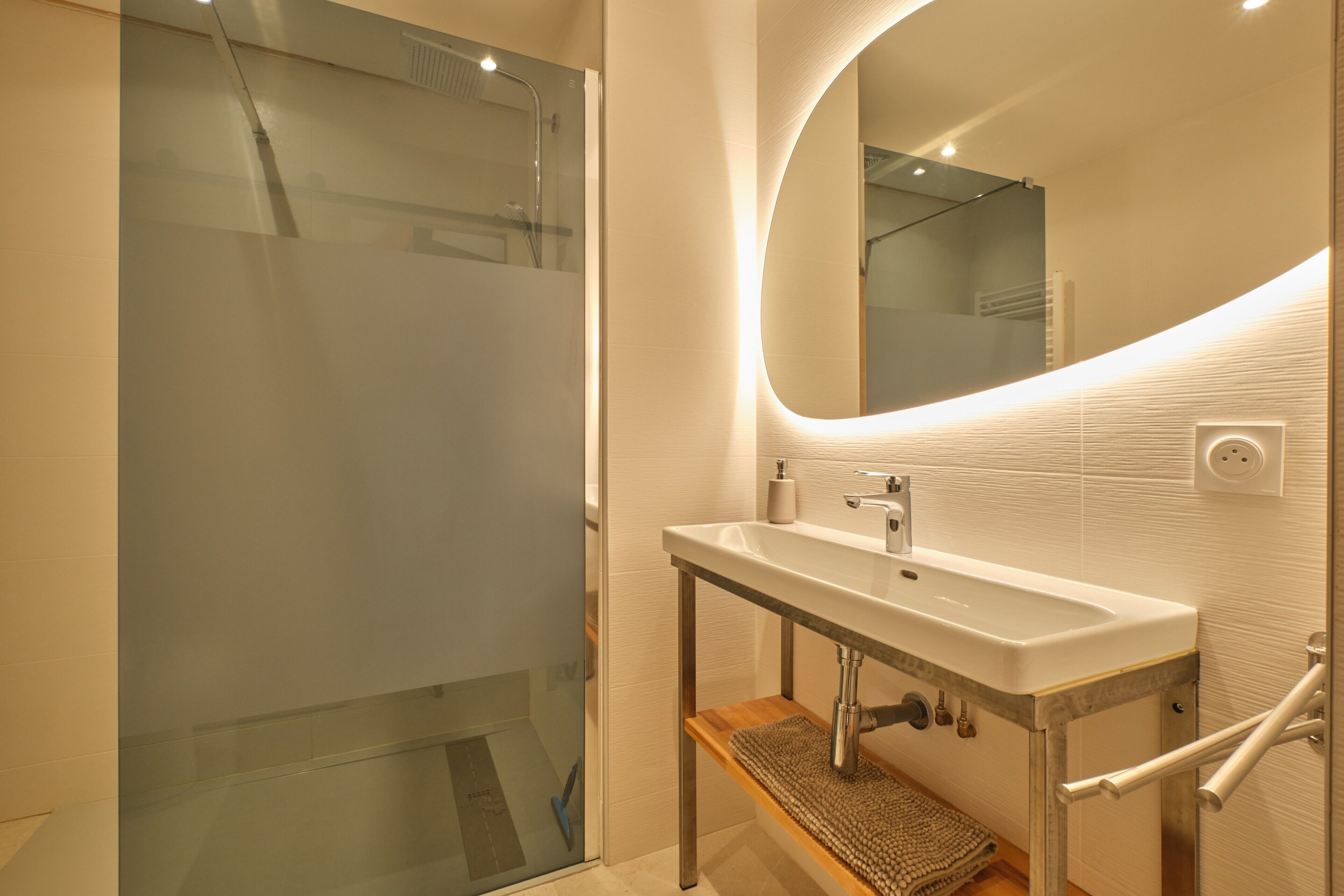 A modern bathroom with a glass shower enclosure, a white sink with a wooden shelf, and a large mirror.