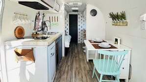 Cocina compacta con lavamanos, estufa y armarios blancos. Cuenta con un comedor con mesa y sillas, además de una pequeña planta en la pared.