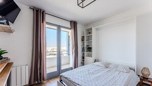 A bedroom with a bed, a sliding glass door to a balcony, a white shelving unit, and a small table with a lamp.