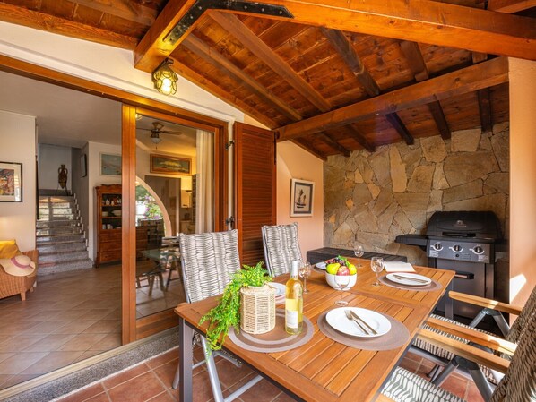 A wooden dining table set with plates, cutlery, and a bottle of wine, with a stone wall and a grill in the background. - Cozy Holiday Home "Casa Oleandro" in Villasimius – Relaxation & Comfort Near the Sea (Villasimius)