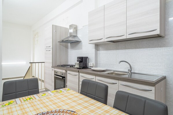 A compact kitchen with a stove, sink, and cabinets, a dining table with chairs, and a staircase leading to another room.